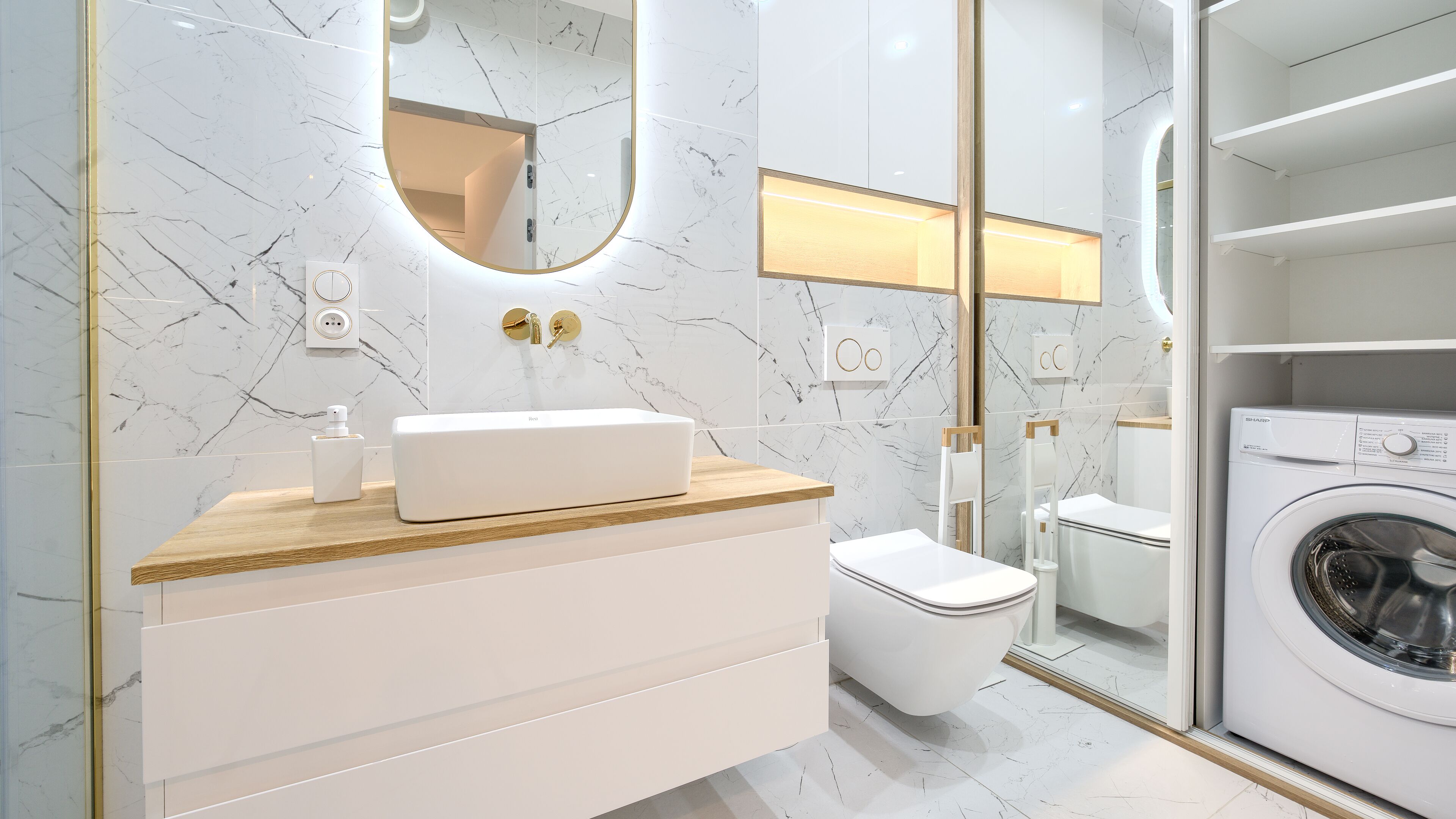 A modern bathroom with a marble backsplash, a white vanity with a gold accent, a toilet, a washing machine, and a large mirror.