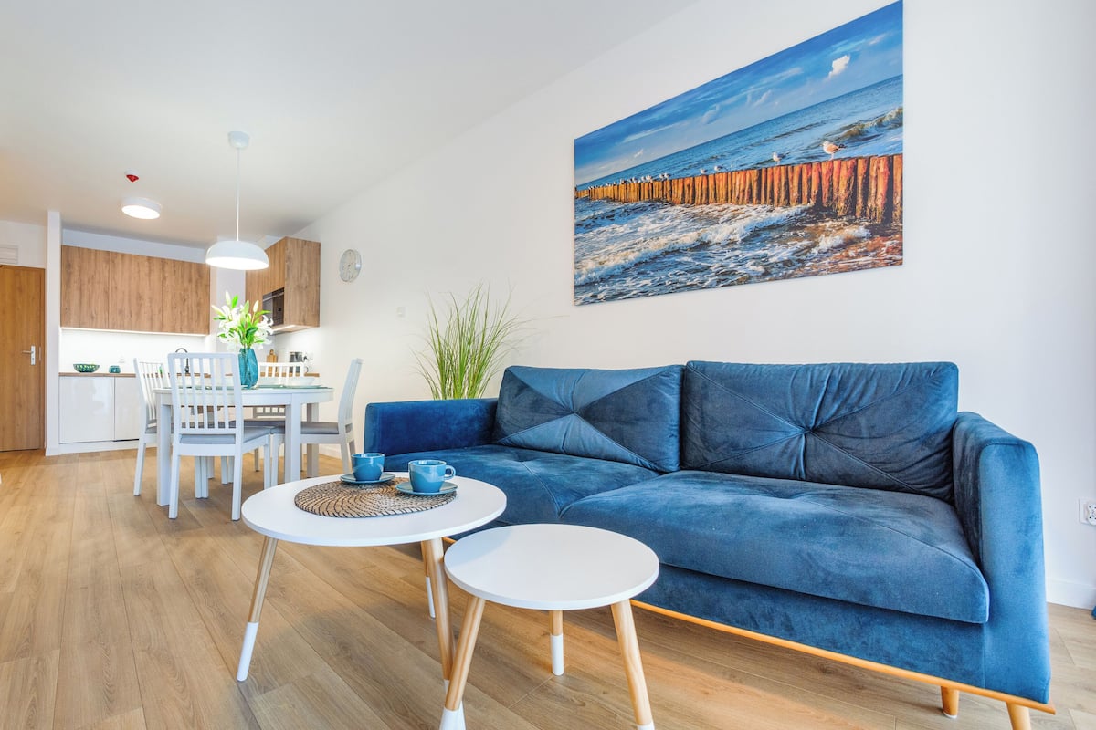 A modern living room with a blue sofa, a dining table with chairs, a kitchen area, and a large painting of a coastal scene.
