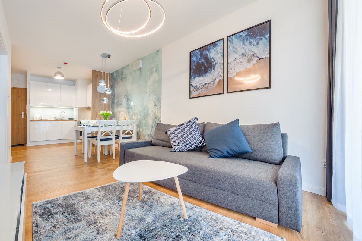 A modern living room with a grey sofa, a small white table, and a rug. There is a kitchen in the background with white cabinets and a dining area.