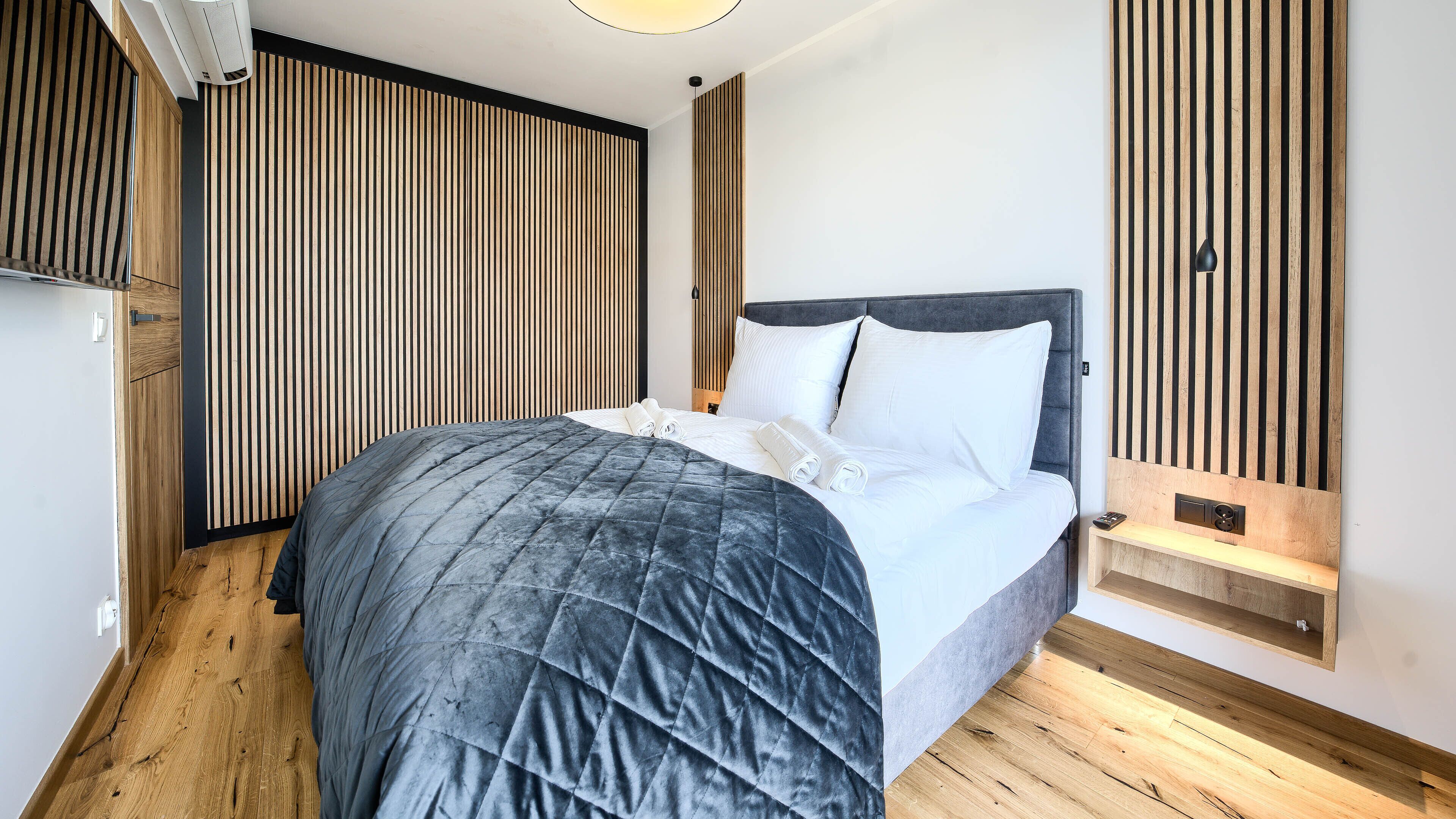A modern bedroom with a large bed, wooden panel wall, and a small bedside table with a lamp.