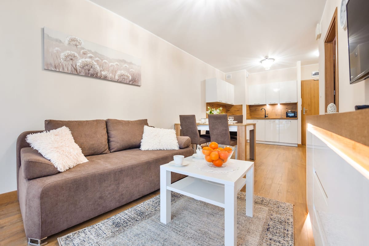 A modern living room with a brown sofa, white cushions, a white coffee table with a bowl of fruit, and a painting on the wall.