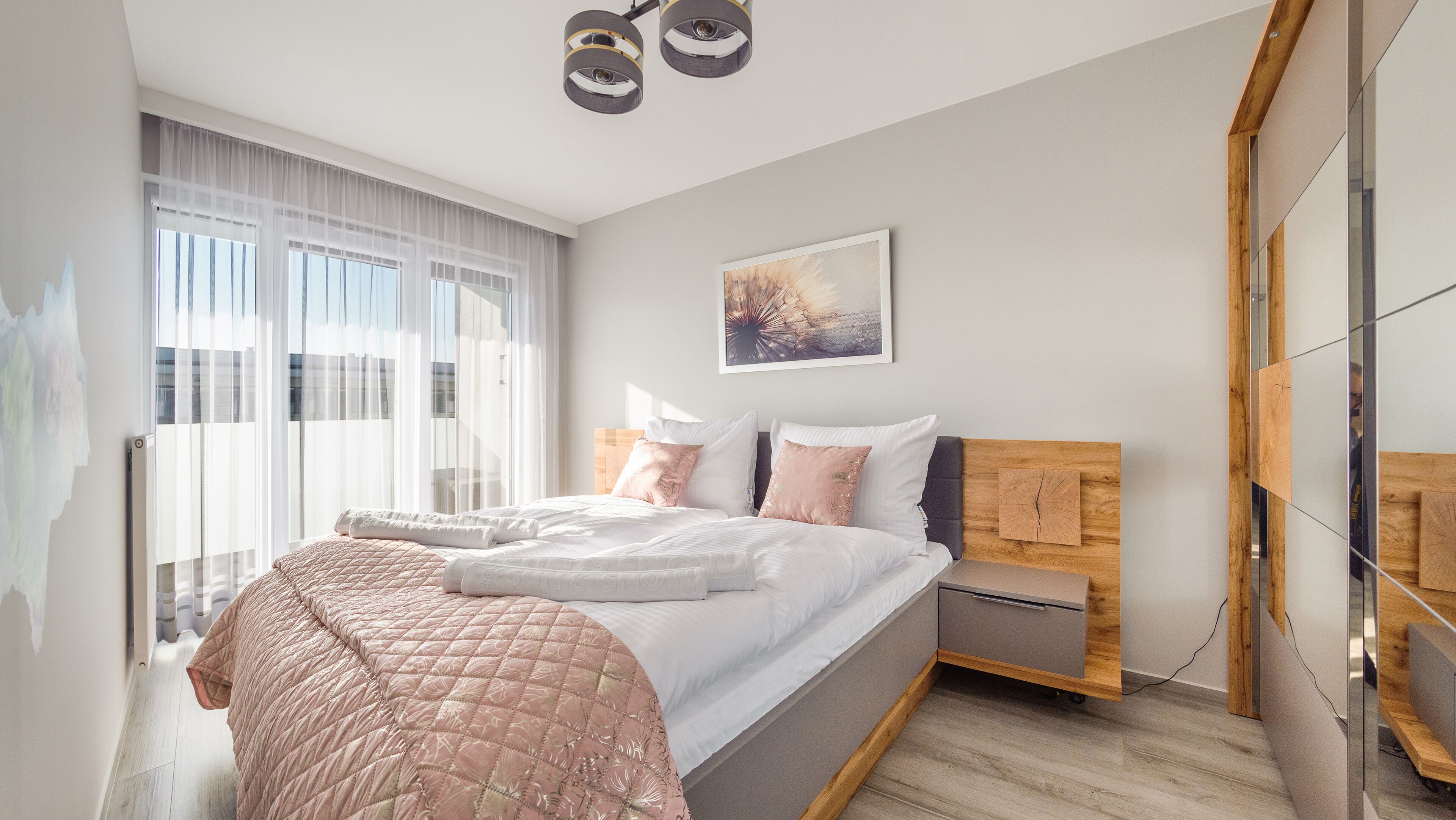 Een moderne slaapkamer met een groot bed, een houten hoofdbord, een nachtkastje en een raam met transparante gordijnen.