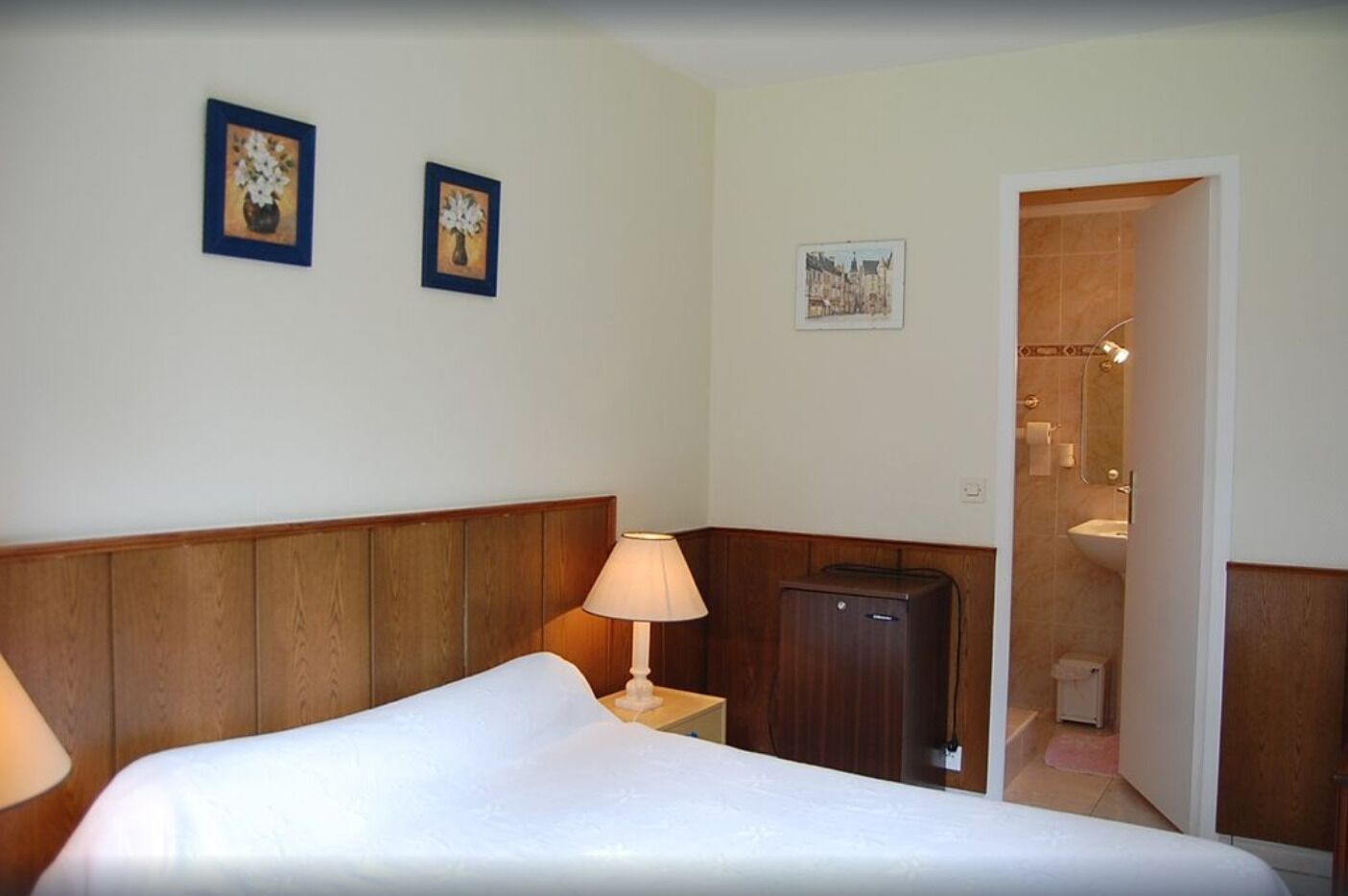 A hotel room with a bed, a nightstand, a lamp, a small cabinet, and a bathroom visible through an open door.