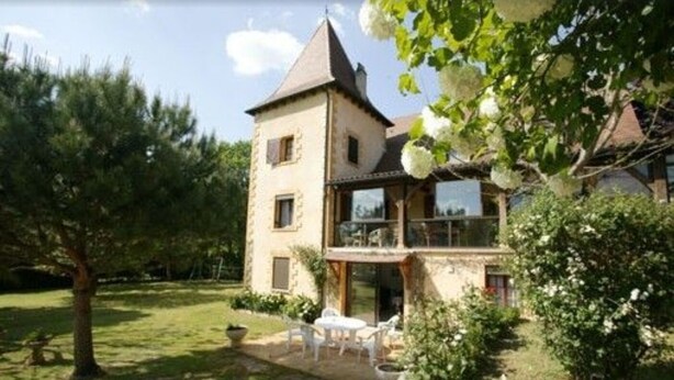 Une maison en pierre à deux étages, dotée d’un balcon, entourée de verdure et d’une pelouse.
