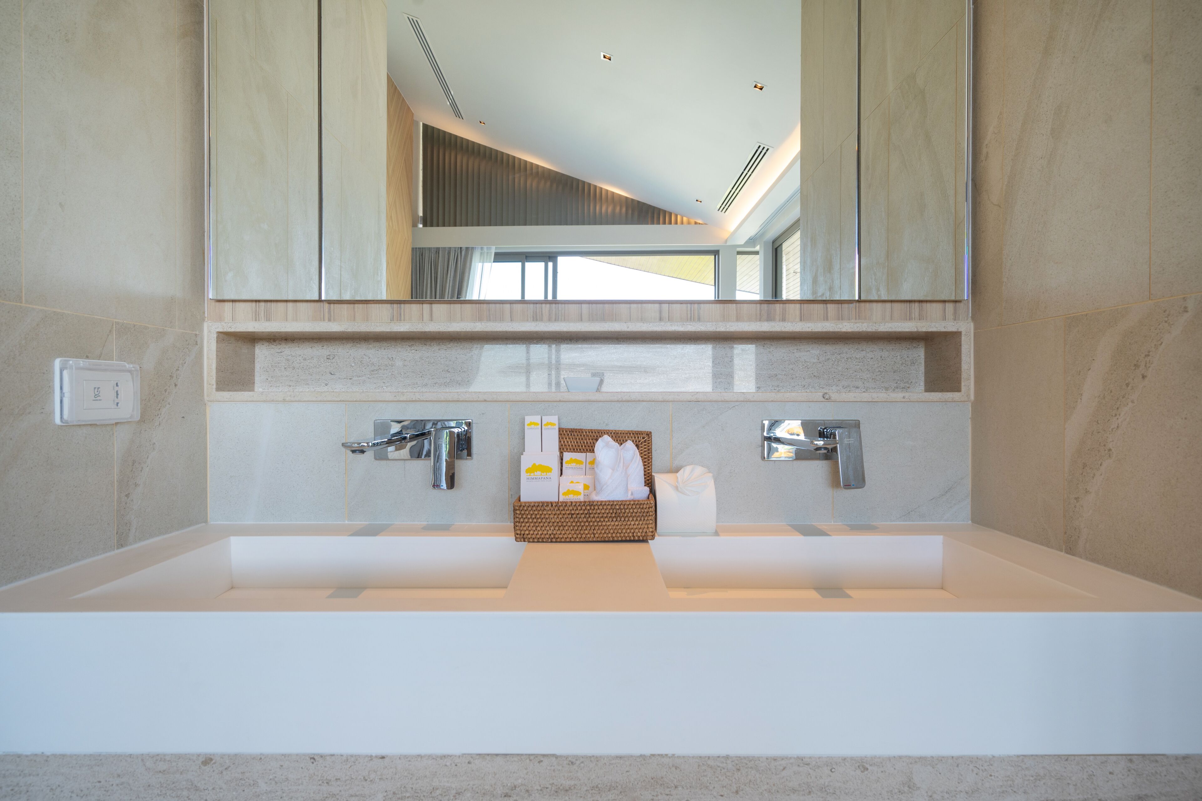 A modern bathroom with a large mirror, two sinks, and a wicker basket with toiletries.