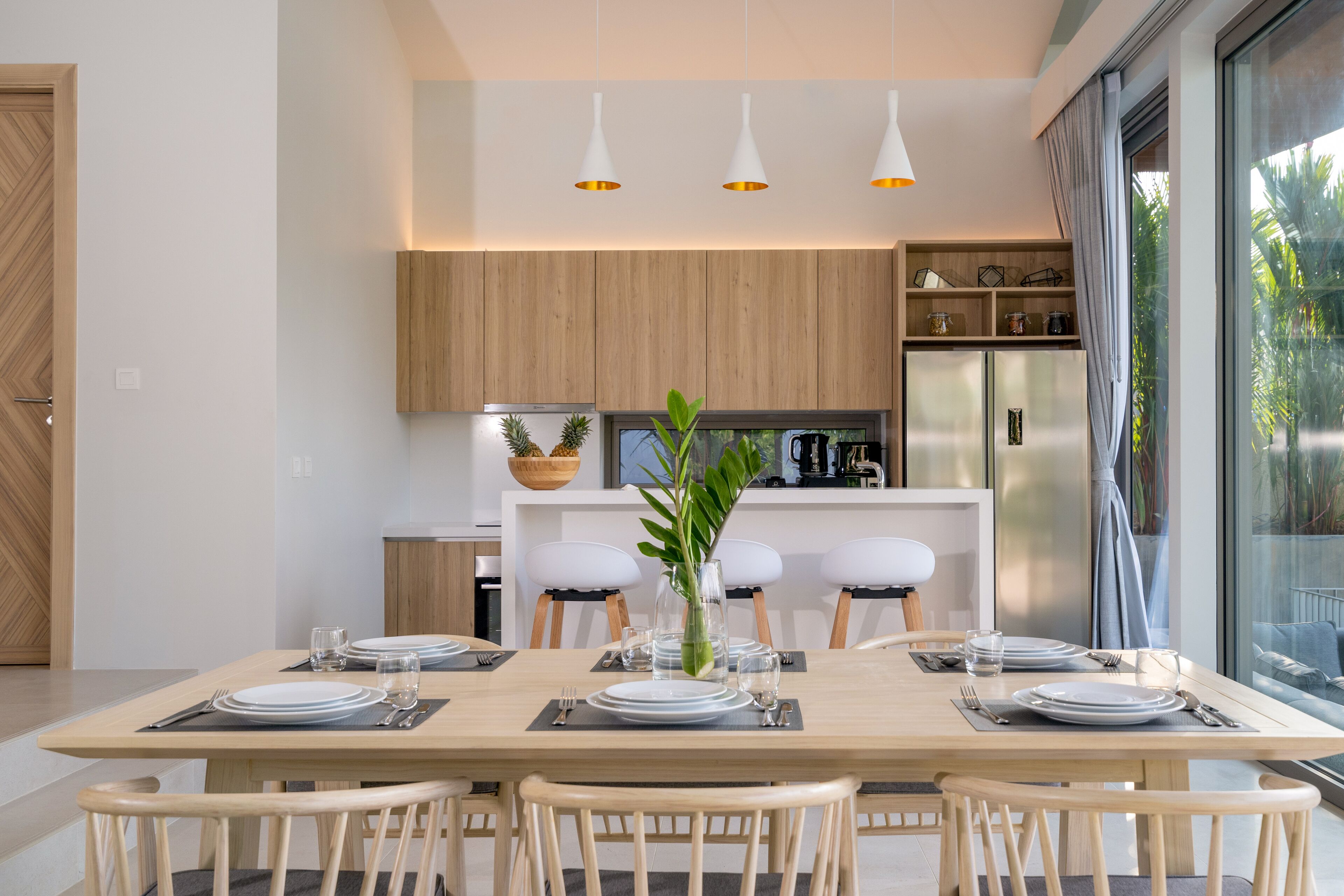 A modern dining area with a wooden table set for four, surrounded by chairs. There are pendant lights above the table, and a kitchen area is visible in the background.