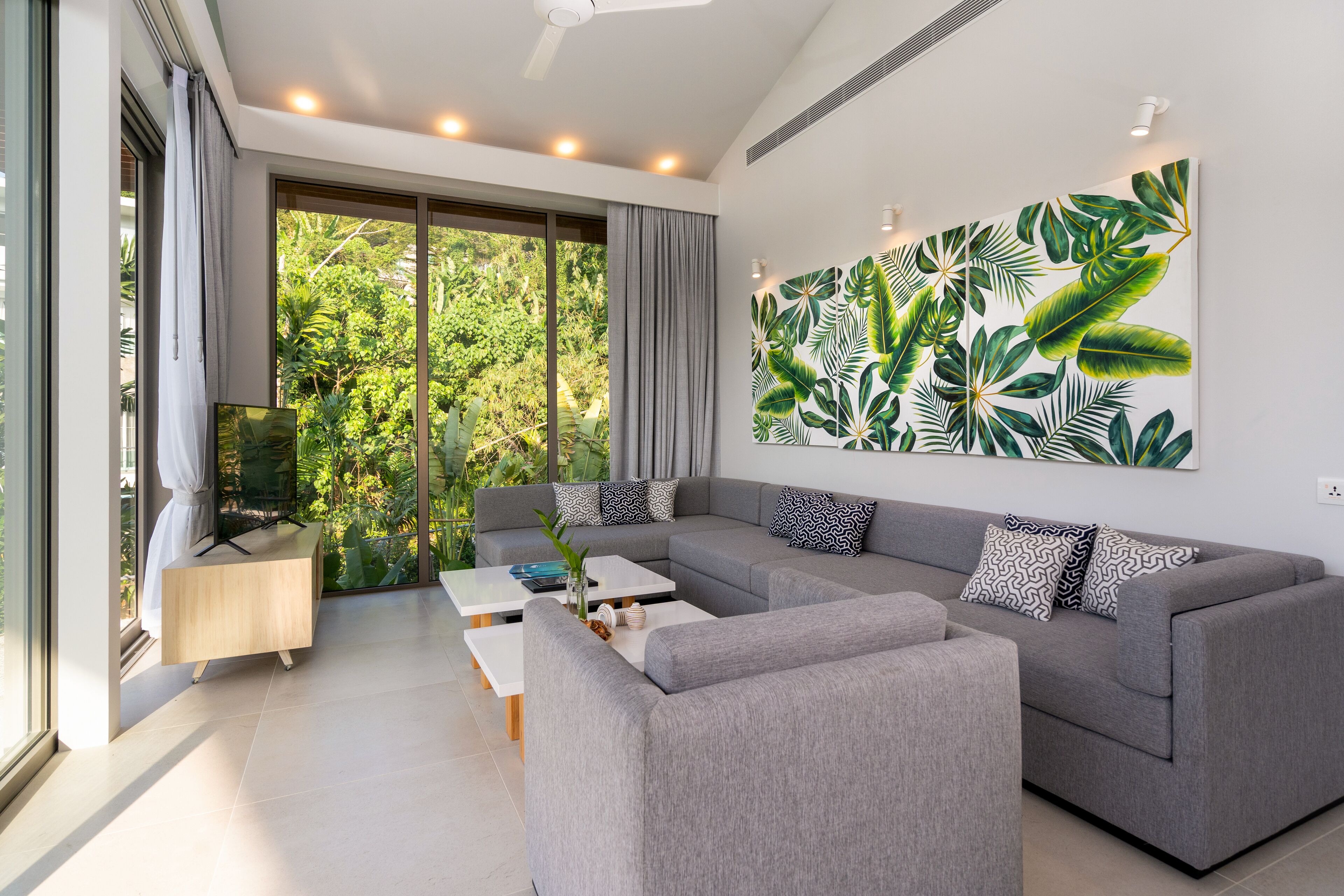 Una sala de estar moderna con un sofá gris, mesas de centro blancas y un mural grande con hojas tropicales en la pared.