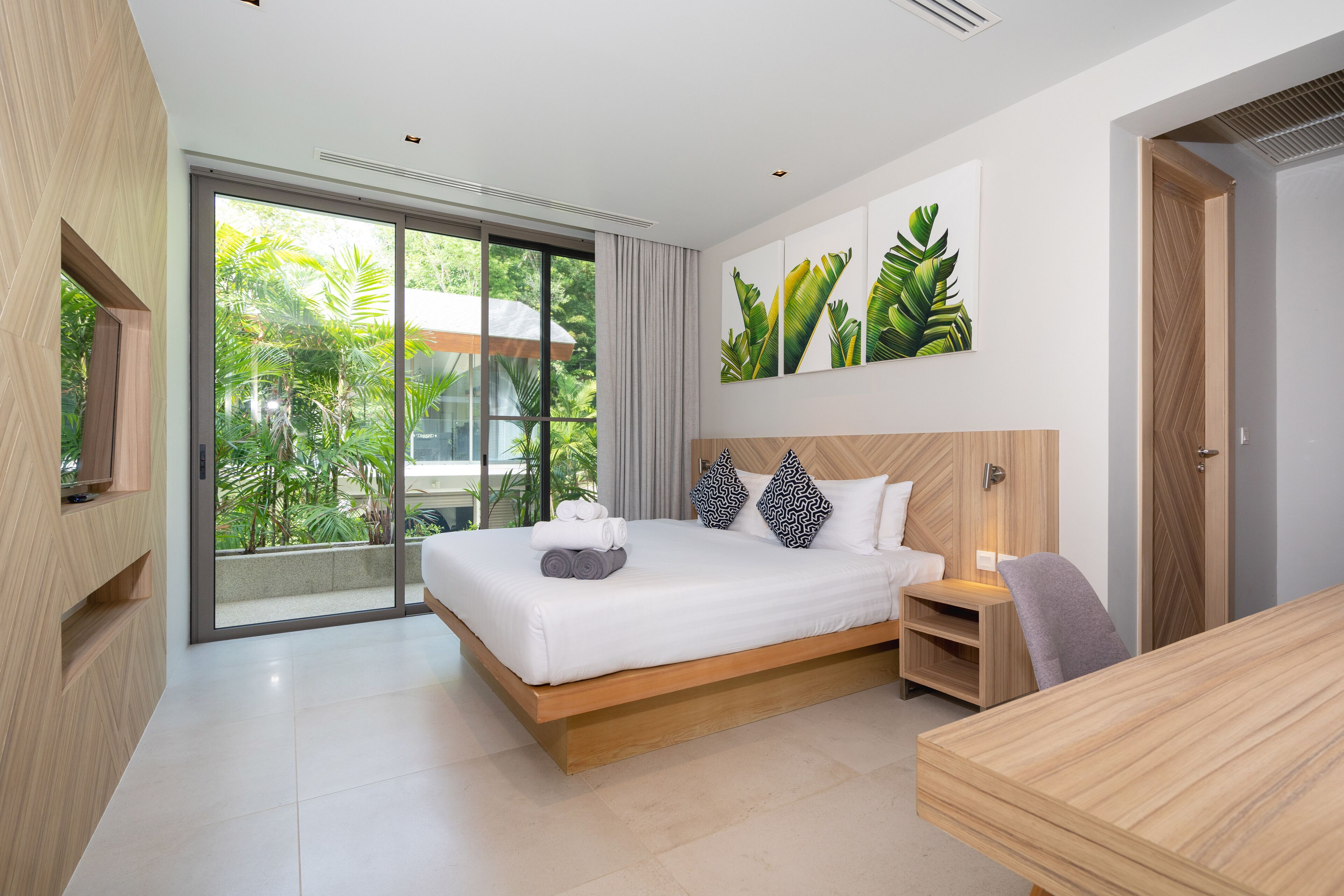 Une chambre d’hôtel moderne dotée d’un grand lit, d’une table de chevet en bois, d’un bureau et d’une porte-fenêtre coulissante donnant sur un balcon agrémenté de plantes.