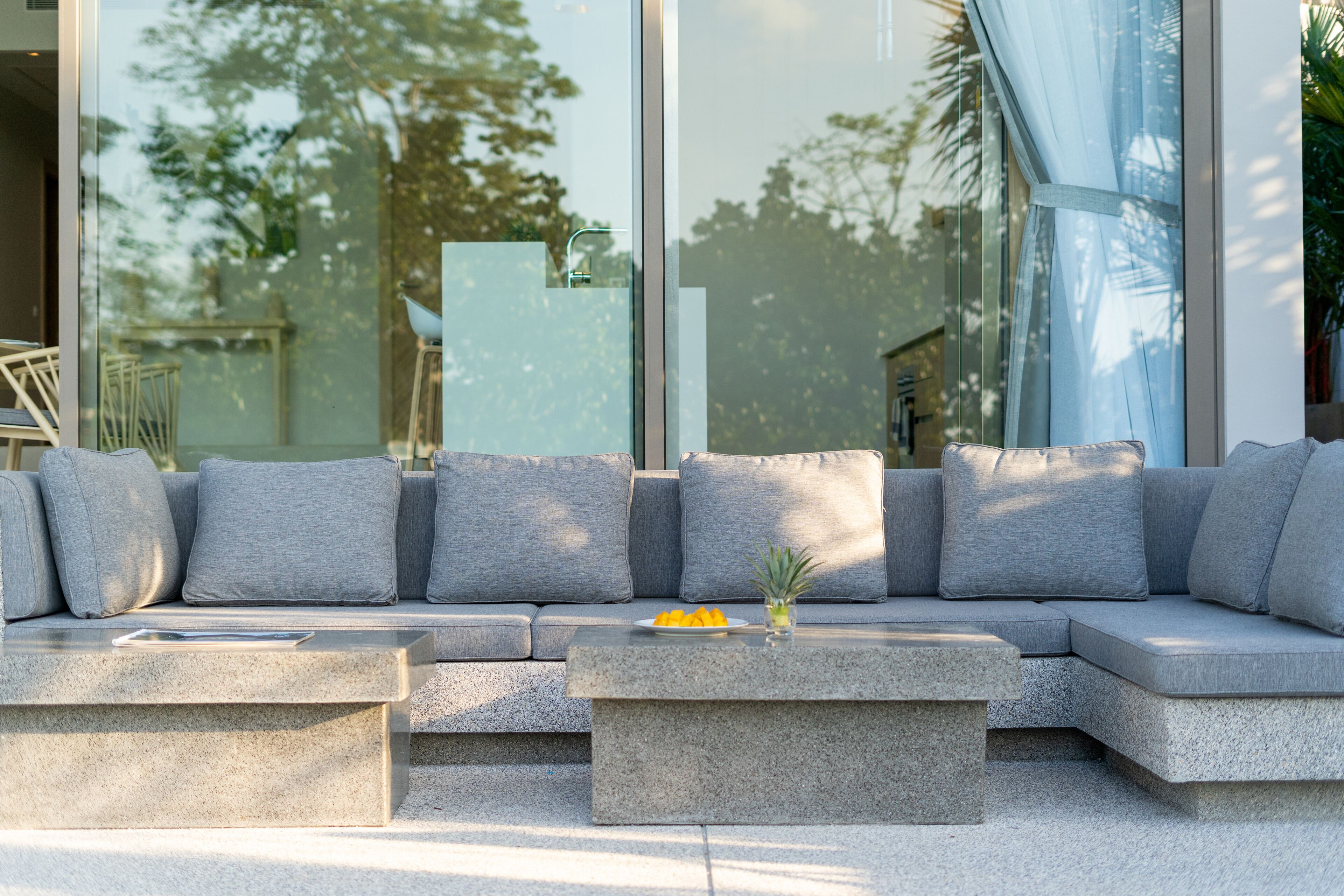 A modern outdoor seating area with a stone bench and a sofa, a small table with a fruit bowl, and a glass wall reflecting trees and a building.