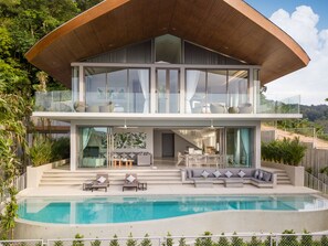 Uma casa moderna de dois andares com uma grande fachada de vidro, piscina e área de refeições com mesas ao ar livre.