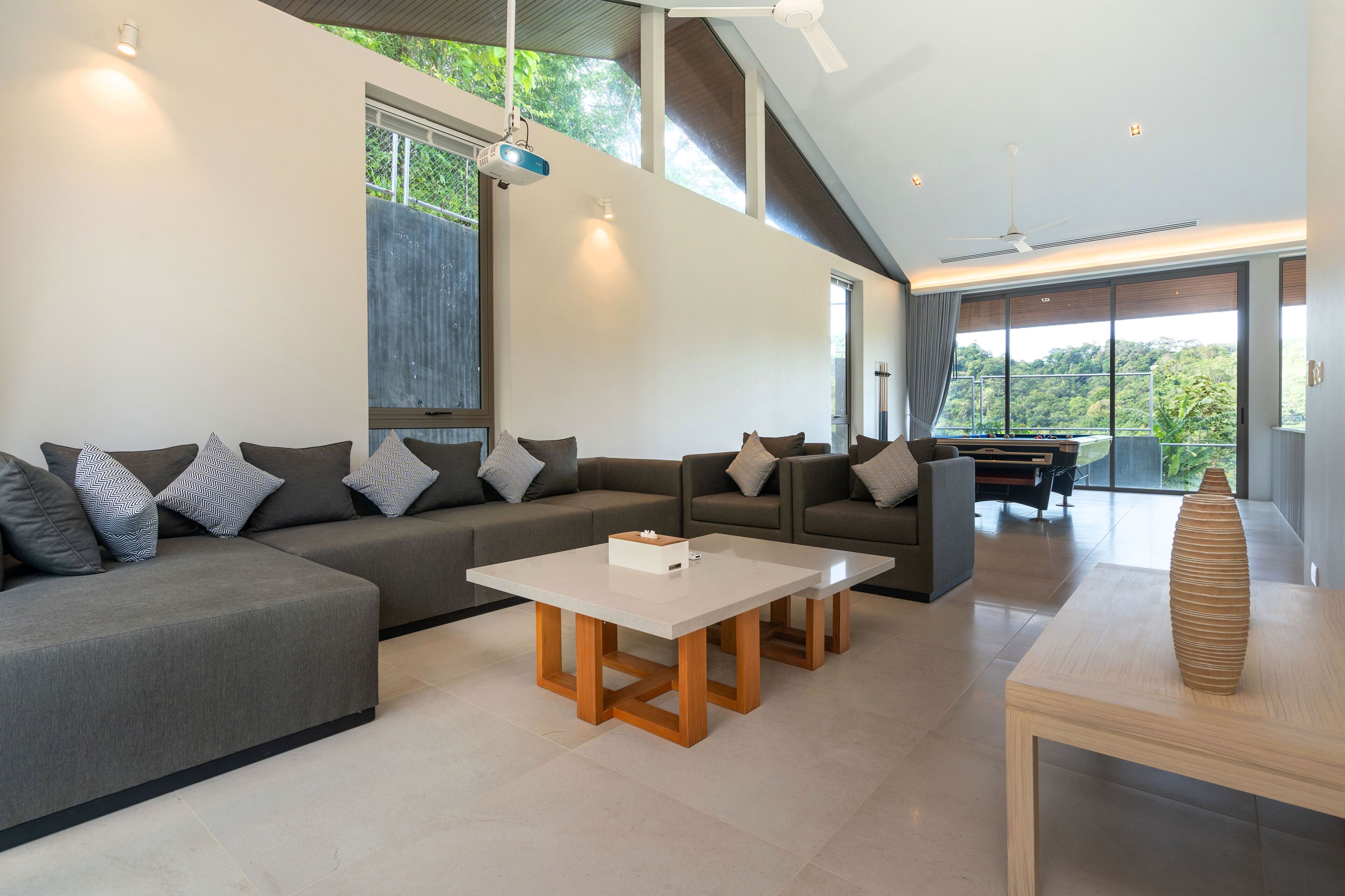 A modern living room with a sectional sofa, a coffee table, and a dining table. There are large windows providing natural light and a view of greenery outside.
