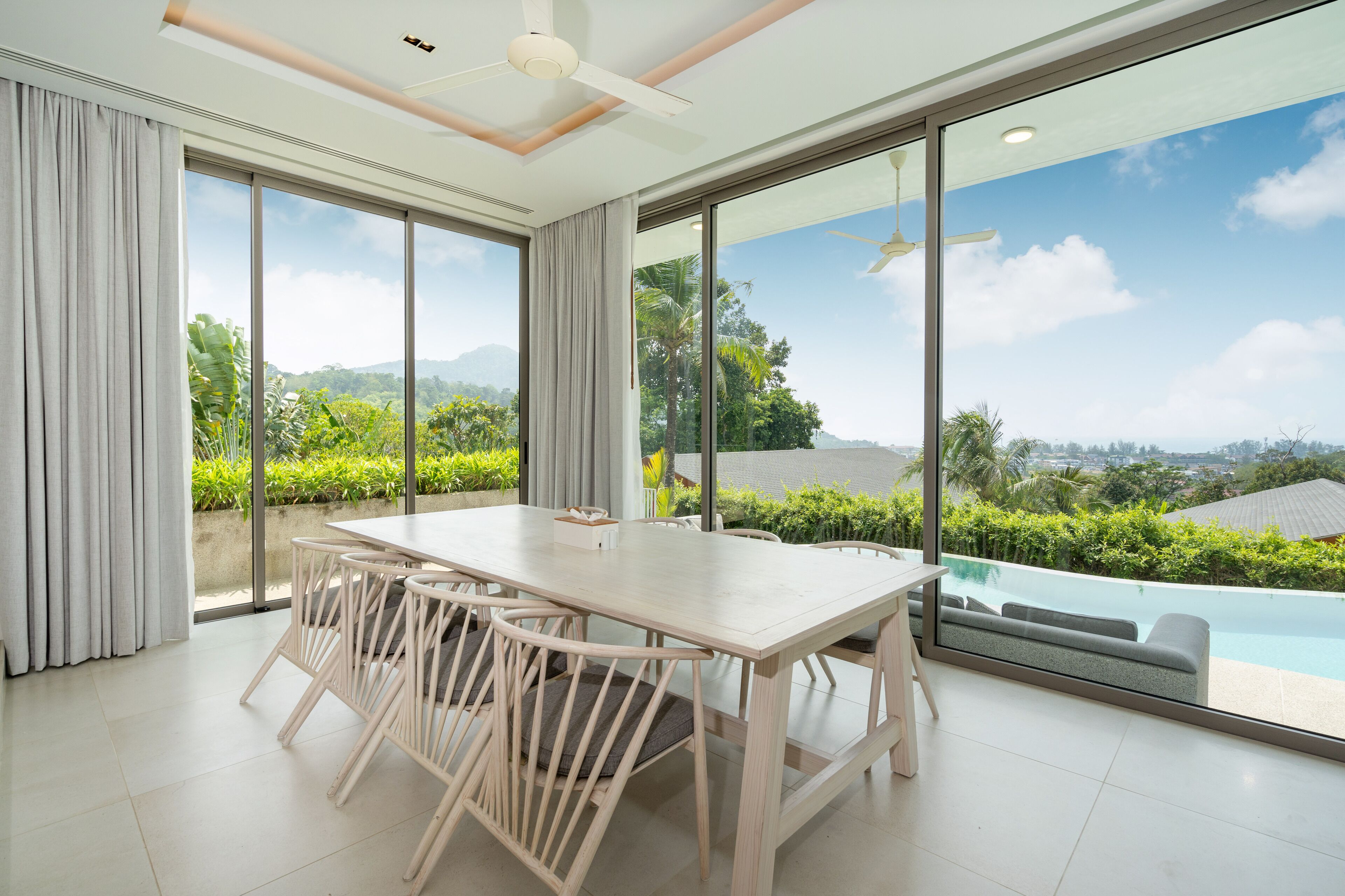 Une pièce spacieuse avec une grande table à manger et des chaises, un ventilateur de plafond et de grandes fenêtres offrant une vue sur une piscine et un espace verdoyant.