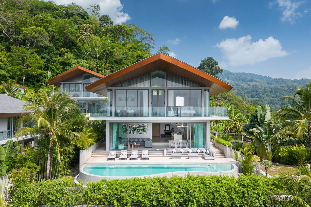 A two-story house with a swimming pool, surrounded by lush greenery and a mountainous backdrop.