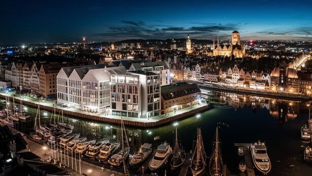A waterfront hotel with a modern glass facade, a marina with boats, and historic buildings in the background.
