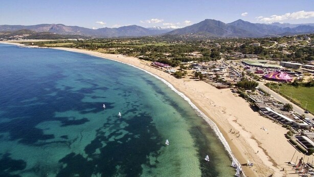 Vista aérea de uma área costeira com praia, águas azul-claras e montanhas ao fundo.