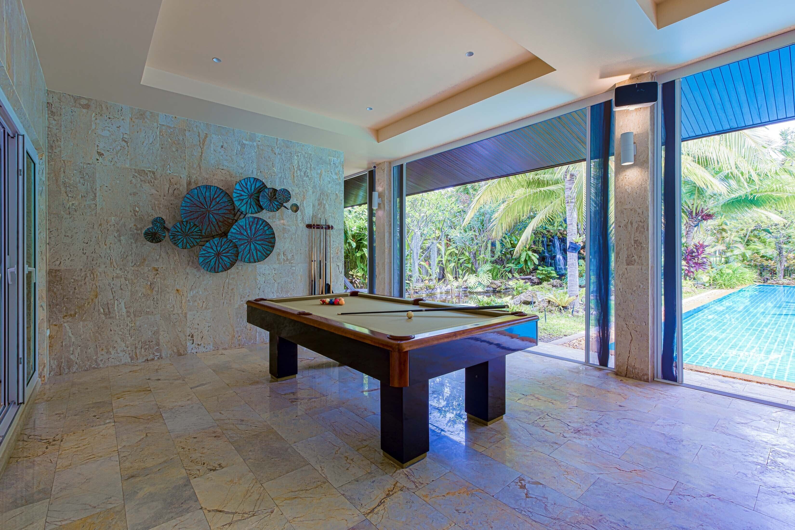 A pool table in a room with a stone wall and large windows overlooking a pool and garden.