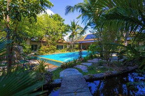 Une piscine entourée d’une végétation luxuriante, un sentier en pierre et un bâtiment doté d’un toit en tuiles.