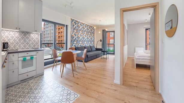 A modern apartment with a kitchen, dining area, and living room. It features a patterned backsplash, wooden flooring, and large windows.