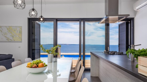 A modern kitchen with a view of the sea, a dining area with a bowl of fruit, and a minimalist design. - Lovely home in Kosljun (Kosljun)