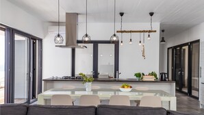A modern kitchen with a central island, pendant lights, and a dining area with a glass table and cushioned chairs. - Lovely home in Kosljun (Kosljun)