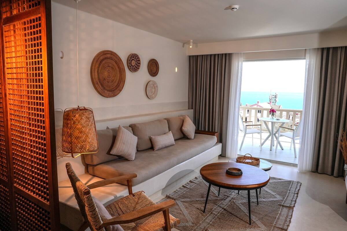 A modern living room with a sofa, a wooden coffee table, wicker chairs, and a balcony with a view of the sea.