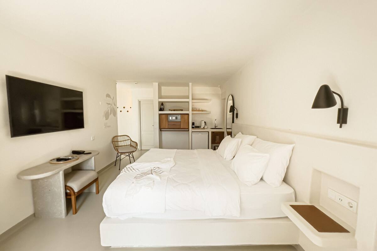 A bedroom with a bed, bedside table, chair, TV, and a kitchenette area.