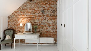 Een kamer met een schuine plafond, een openliggende bakstenen muur, een witte radiator, een houten stoel, een wit bureau met een lamp en een spiegel.
