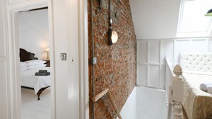 Een slaapkamer met een bakstenen muur, een bed met wit beddengoed, een nachtkastje met een lamp en een raam met uitzicht op buiten.