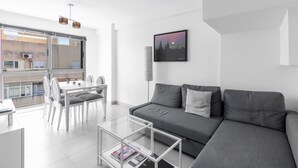 Ein modernes Wohnzimmer mit einer grauen Couch, einem Glastisch und einem Essbereich mit weißen Stühlen und einem Tisch.