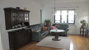 A living room with a dark wooden cabinet, a green sofa, a round coffee table, and a large window with a view of buildings. - Apartment Silbermöwe Object-ID 121011 (Waren (Müritz))