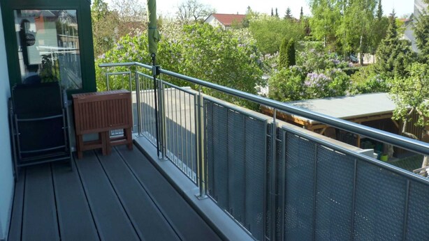 A balcony with a wooden bench, metal railing, and a view of a garden with trees and flowers. - Apartment Silbermöwe Object-ID 121011 (Waren (Müritz))