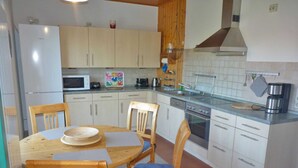 A compact kitchen with white cabinets, a microwave, a stove, and a sink. There are four wooden chairs with blue cushions around a small round table. - Apartment Silbermöwe Object-ID 121011 (Waren (Müritz))