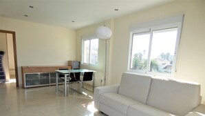 A modern living room with a white sofa, a glass-top dining table with two chairs, a small TV, and a window with a view of palm trees.