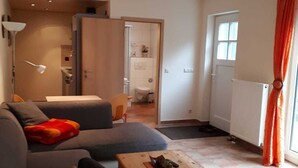 A modern living room with a grey sofa, a wooden coffee table, and a wooden chair. There is a door leading to a bathroom and a window with orange curtains. - Apartment on Gregoriusberg Object-ID 121158 (Waren (Müritz))