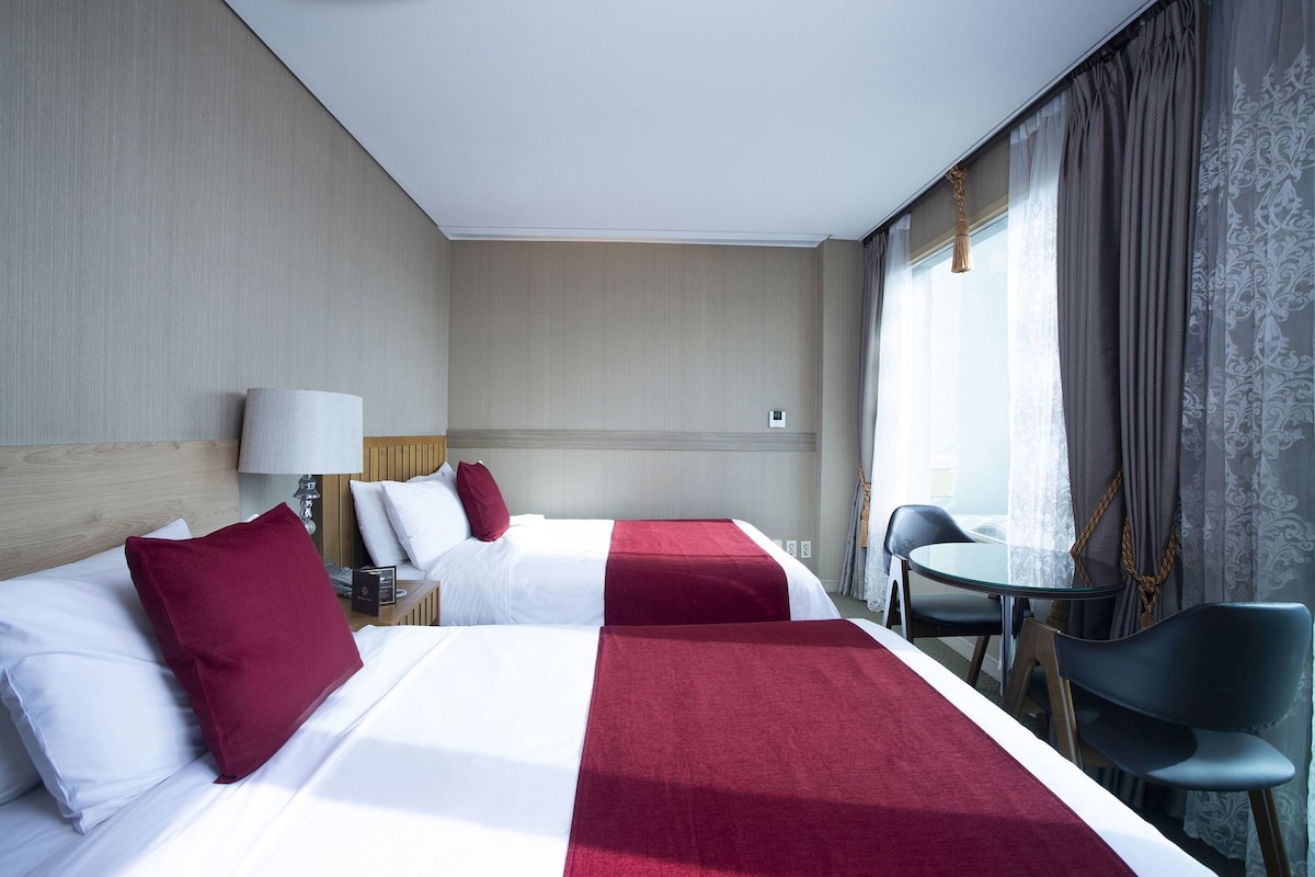 Una habitación de hotel con dos camas, un escritorio, una silla y un ventanal con cortinas.