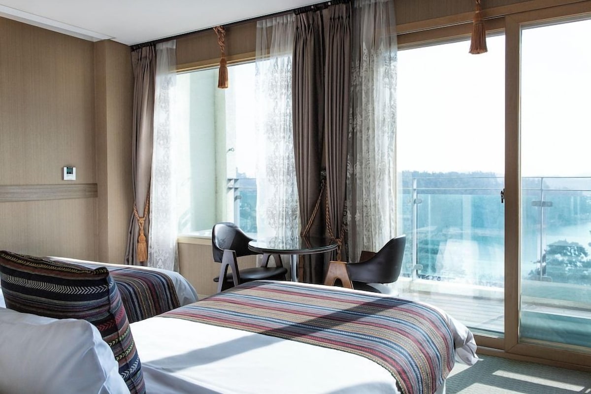 Habitación de hotel con una cama, dos sillas, una mesita pequeña y vistas a un paisaje urbano a través de amplios ventanales.
