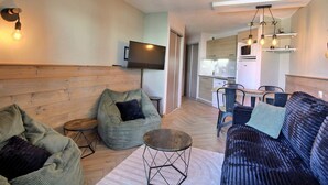 A compact living space with a sofa, bean bag, a small round table, a flat-screen TV mounted on the wall, a kitchenette, and a dining area with chairs.