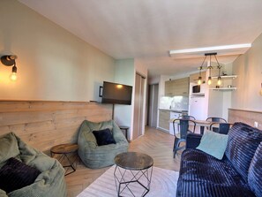 A compact living space with a sofa, bean bag, a small round table, a flat-screen TV mounted on the wall, a kitchenette, and a dining area with chairs.