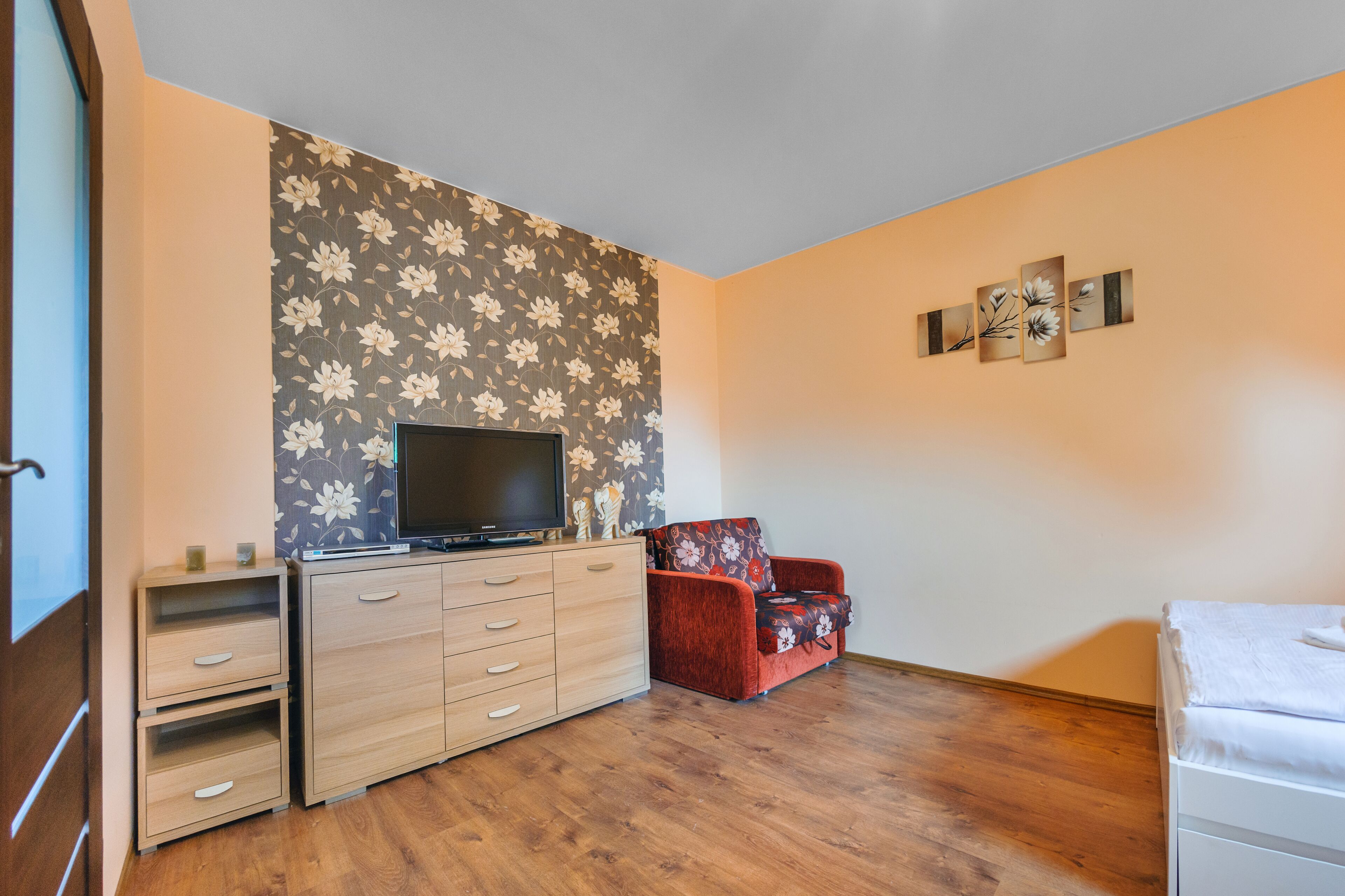 A room with a wooden floor, a TV on a wooden cabinet, a red floral sofa, and a white bed.