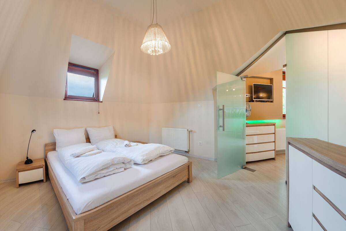 Une chambre moderne avec un lit en bois, une table de chevet, un luminaire suspendu et une vue sur la cuisine par une porte ouverte.
