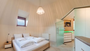 A modern bedroom with a wooden bed, a bedside table, a hanging light fixture, and a view of the kitchen through an open door.