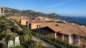 A coastal residential area with terracotta-roofed houses, a clear view of the sea, and a mountain in the background.