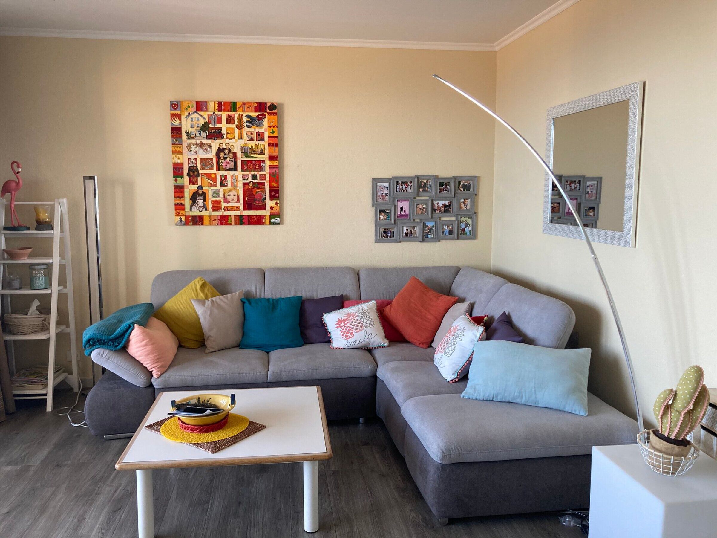 Ein Wohnzimmer mit grauem Sofa, bunten Kissen, einem weißen Couchtisch, einer Wandcollage und einem Spiegel.