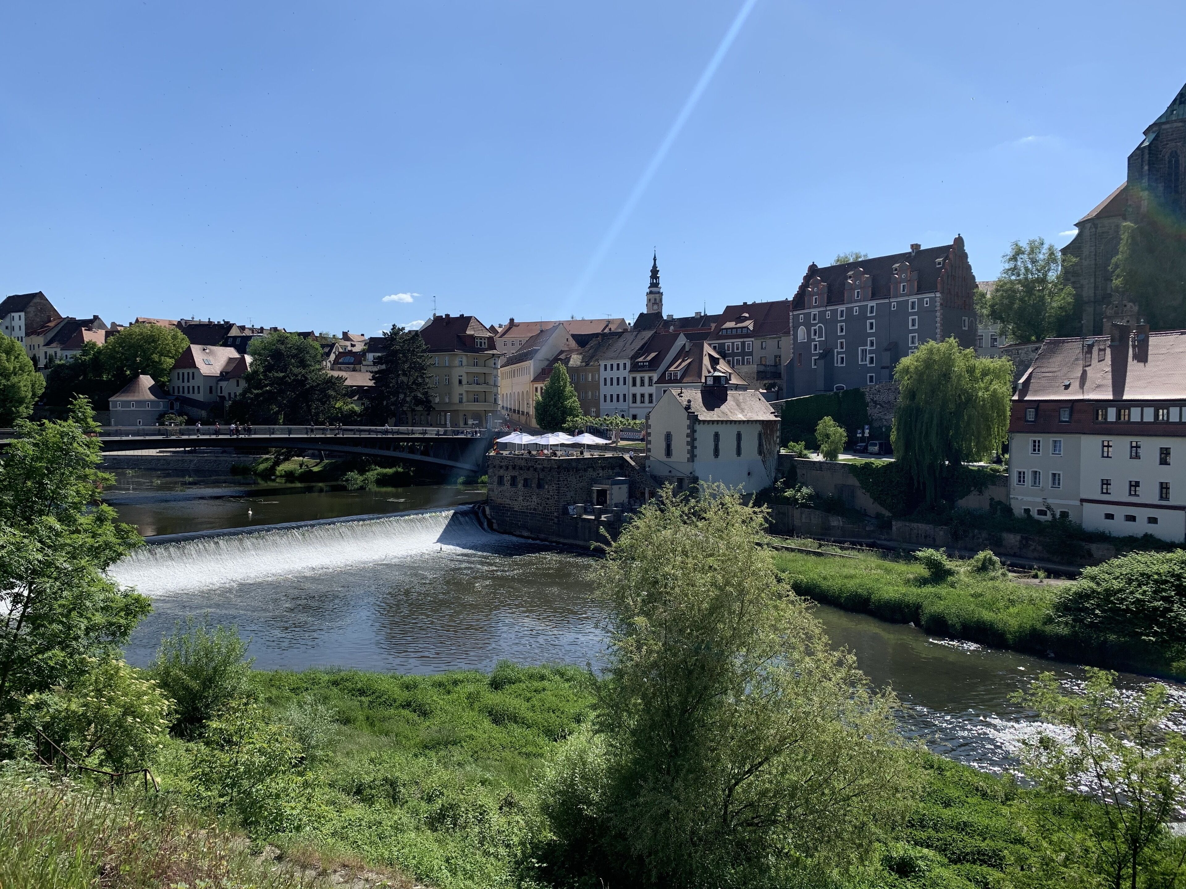 Vista a la orilla del río de un pueblo con edificios históricos, un puente y una cascada.