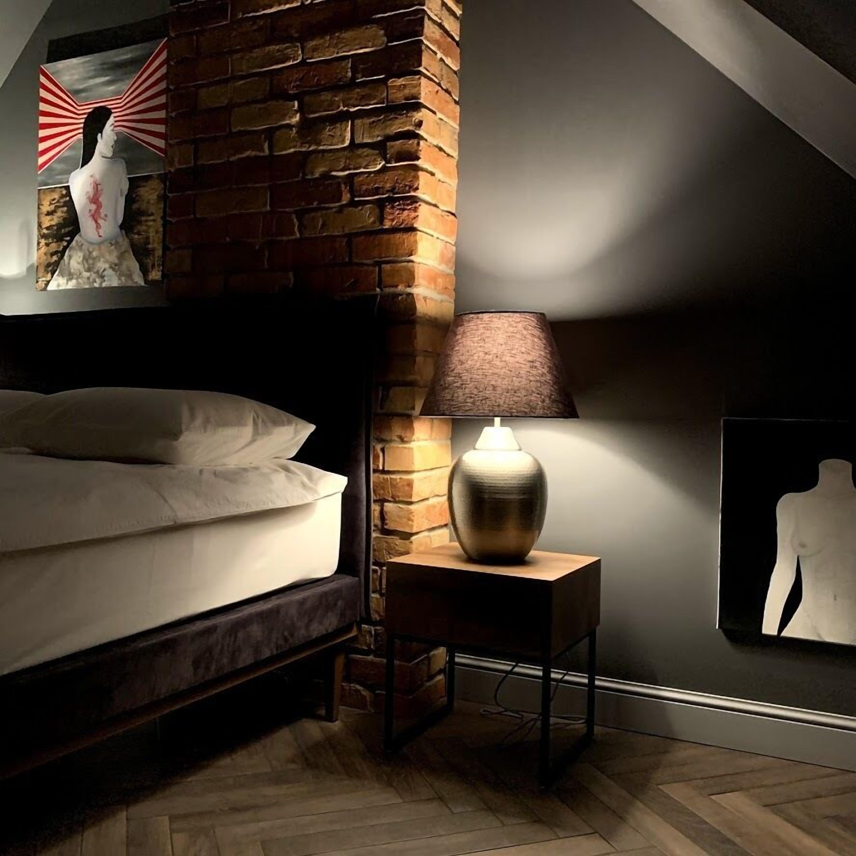 A bedroom with a brick wall, a bed, a lamp on a nightstand, and artwork on the walls.