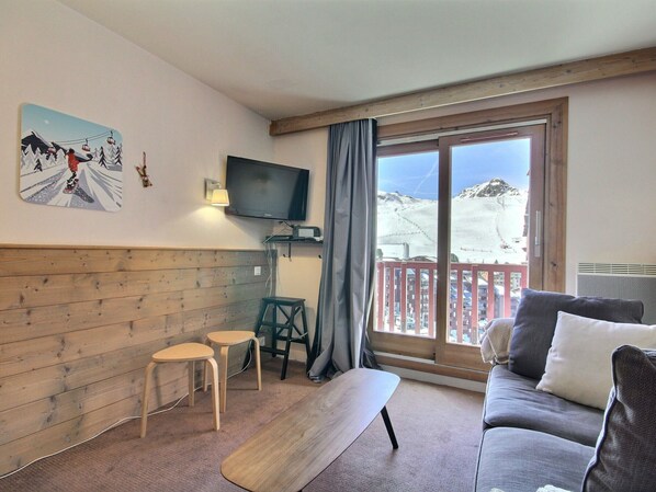 Quarto com parede de madeira, TV, um banco, um sofá e vista para uma montanha nevada.