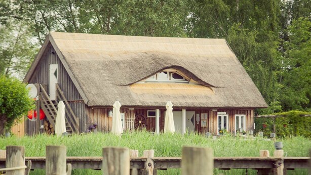 A rustic wooden house with a thatched roof, situated on stilts over water, surrounded by lush greenery.