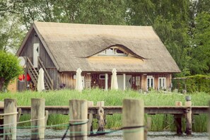 A rustic wooden house with a thatched roof, situated on stilts over water, surrounded by lush greenery. - 2. Backbordkajüte - Kojenhaus (Krummin)