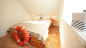 Um quarto pequeno e acolhedor com uma cama de madeira, um salva-vidas no chão e uma mala no parapeito da janela.