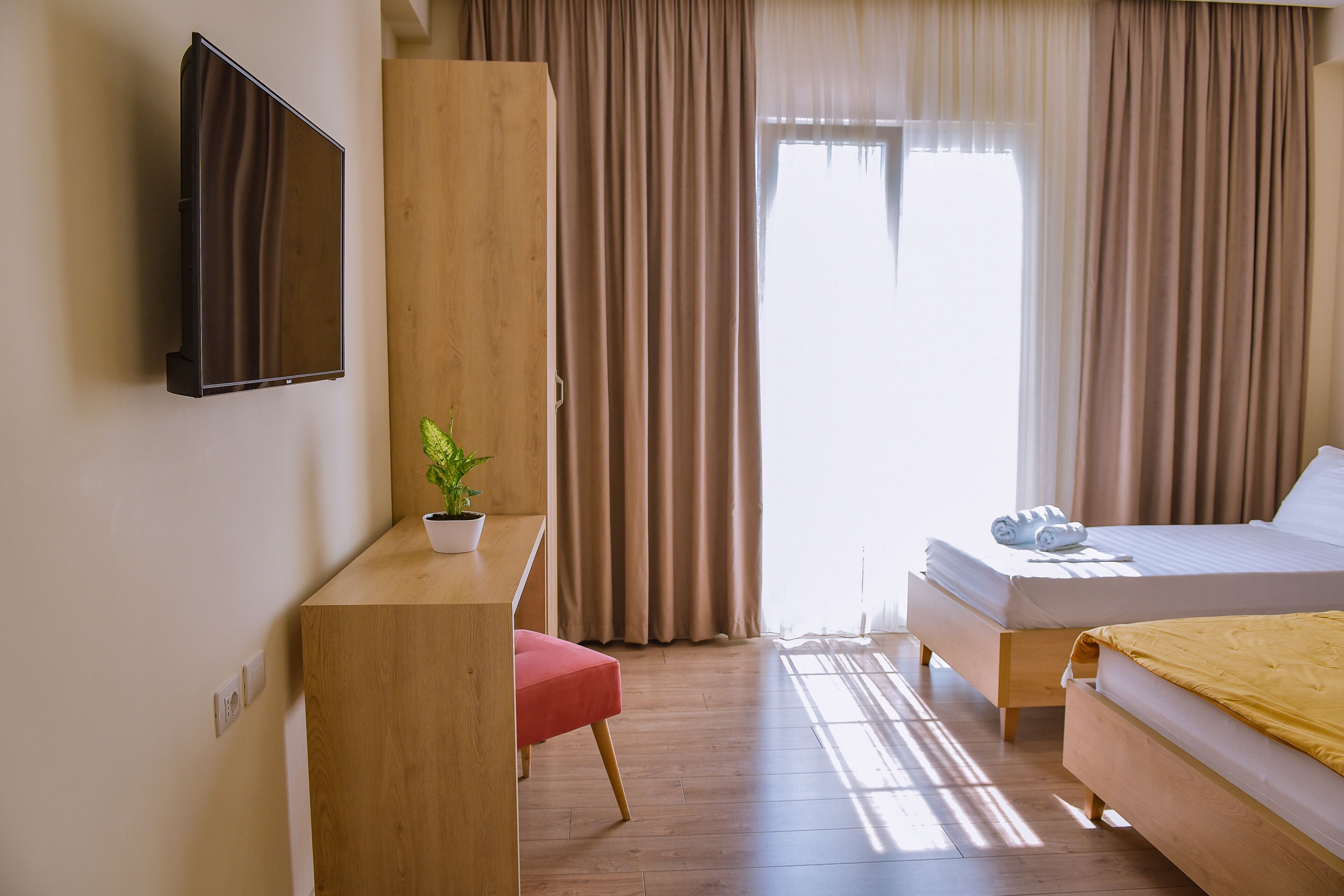 A hotel room with a bed, a desk with a plant, a TV mounted on the wall, and a window with curtains.